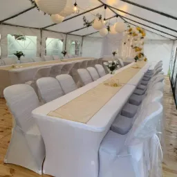 Uroczysty namiot z długimi stołami przykrytymi białymi obrusami i bieżnikami w odcieniu ecru, krzesła w białych pokrowcach, papierowe lampiony i girlanda z balonów w kolorach bieli i złota.