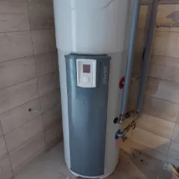 Bojler Atlantic z podłączonymi rurami, widoczne przyłącza hydrauliczne, montaż w pomieszczeniu wyłożonym płytkami imitującymi drewno, na podłodze drobne zabrudzenia.