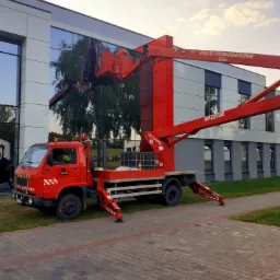 Czerwony podnośnik koszowy ZAMLIFT na tle nowoczesnego budynku z lustrzanymi oknami, dwóch pracowników w koszu na wysokości podczas pracy.