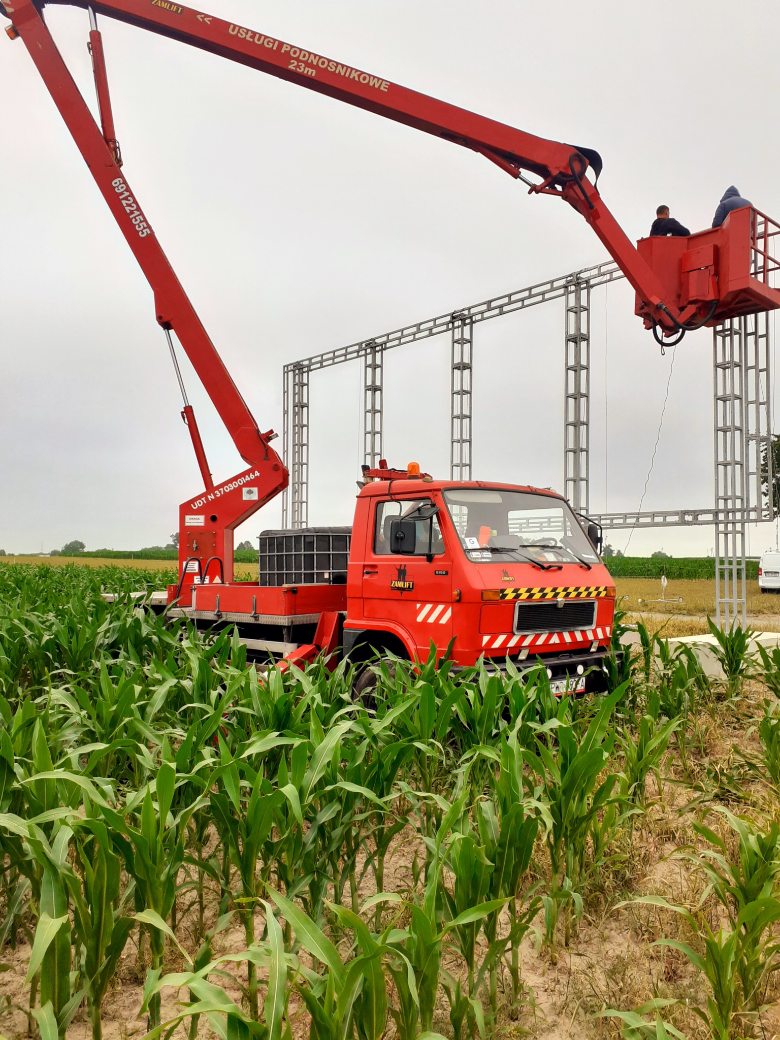 Montaż konstrukcji billboardu za pomocą czerwonego podnośnika koszowego na tle pola kukurydzy.