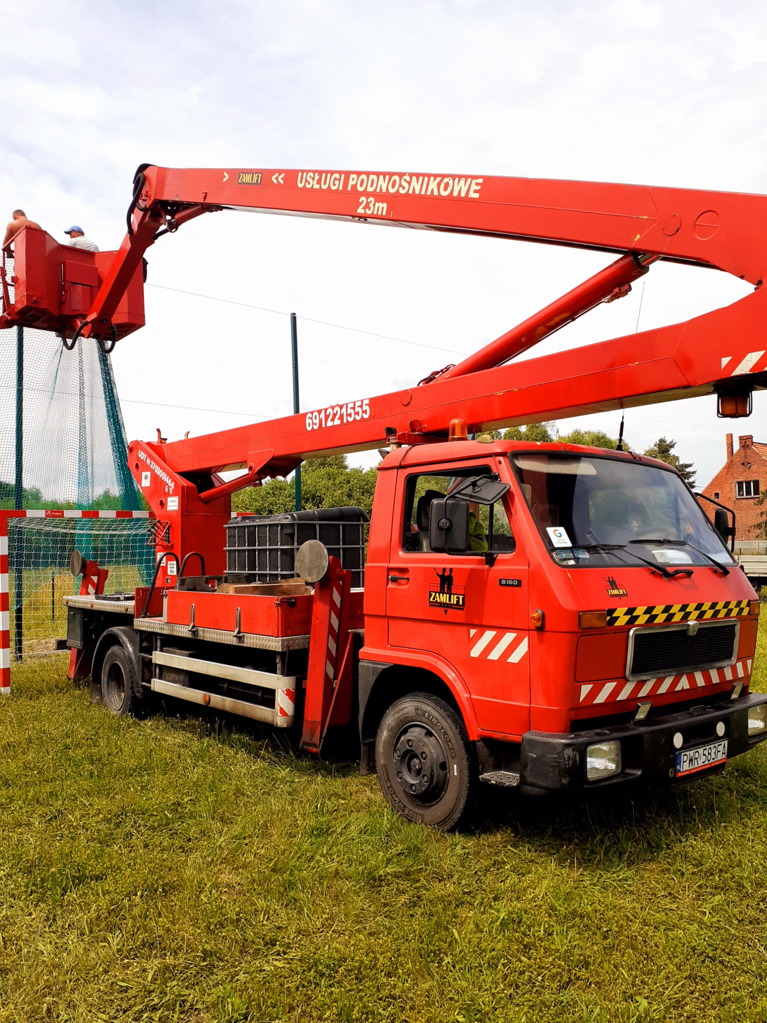 Czerwony podnośnik koszowy ZAMLIFT B150 na trawiastym terenie, z dwoma pracownikami w koszu, ustawiony obok boiska z siatką.
