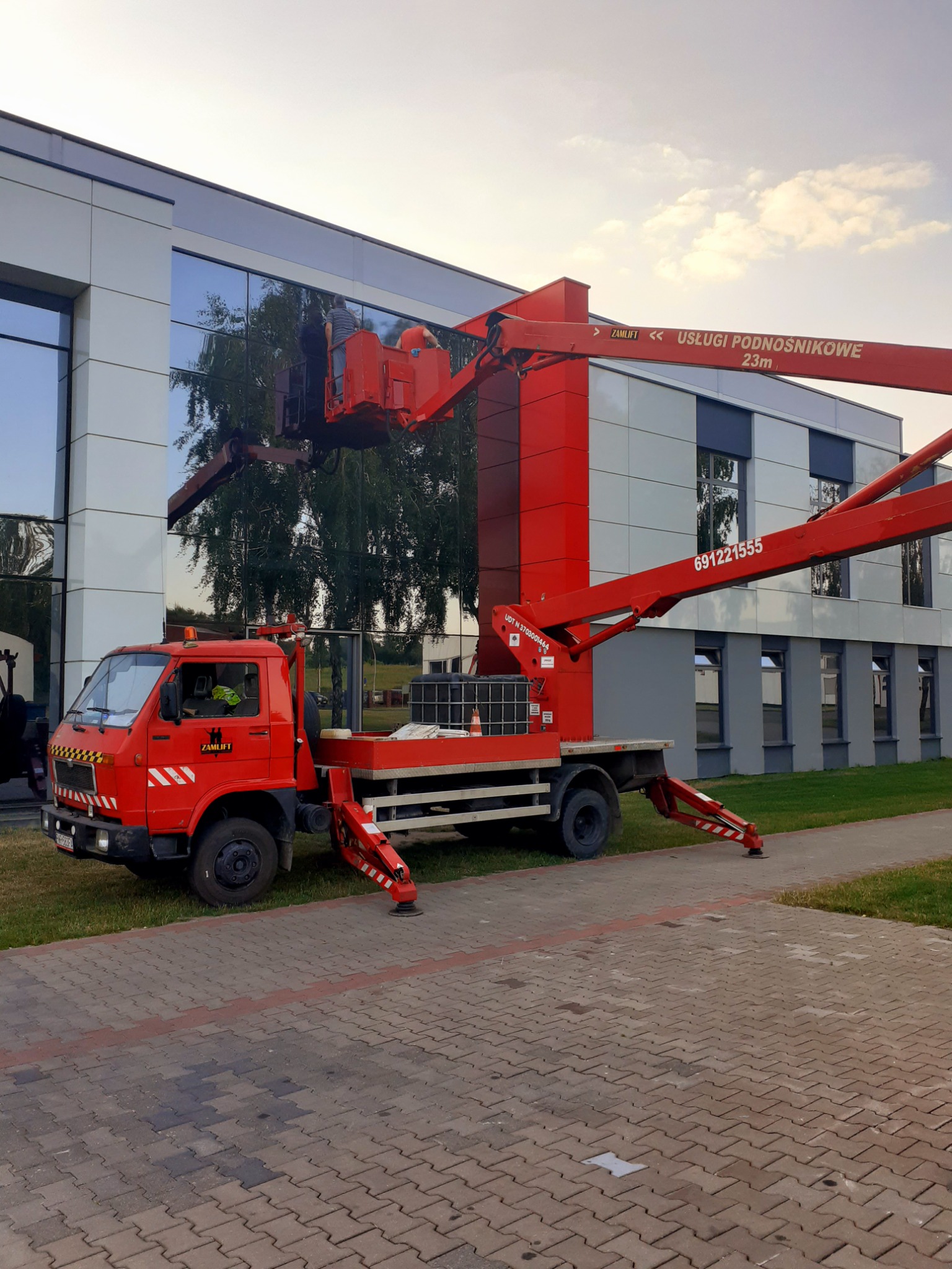 Czerwony podnośnik koszowy ZAMLIFT na tle nowoczesnego budynku z lustrzanymi oknami, dwóch pracowników w koszu na wysokości podczas pracy.