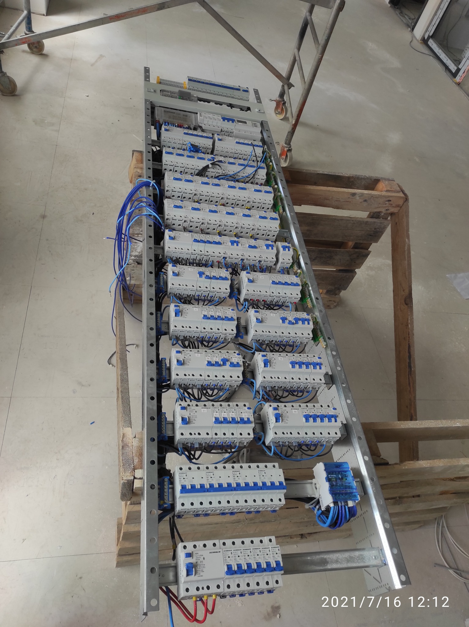 Panel elektryczny z zamontowanymi bezpiecznikami i okablowaniem, ustawiony na drewnianej palecie w pomieszczeniu w trakcie instalacji, widoczne rusztowanie w tle.