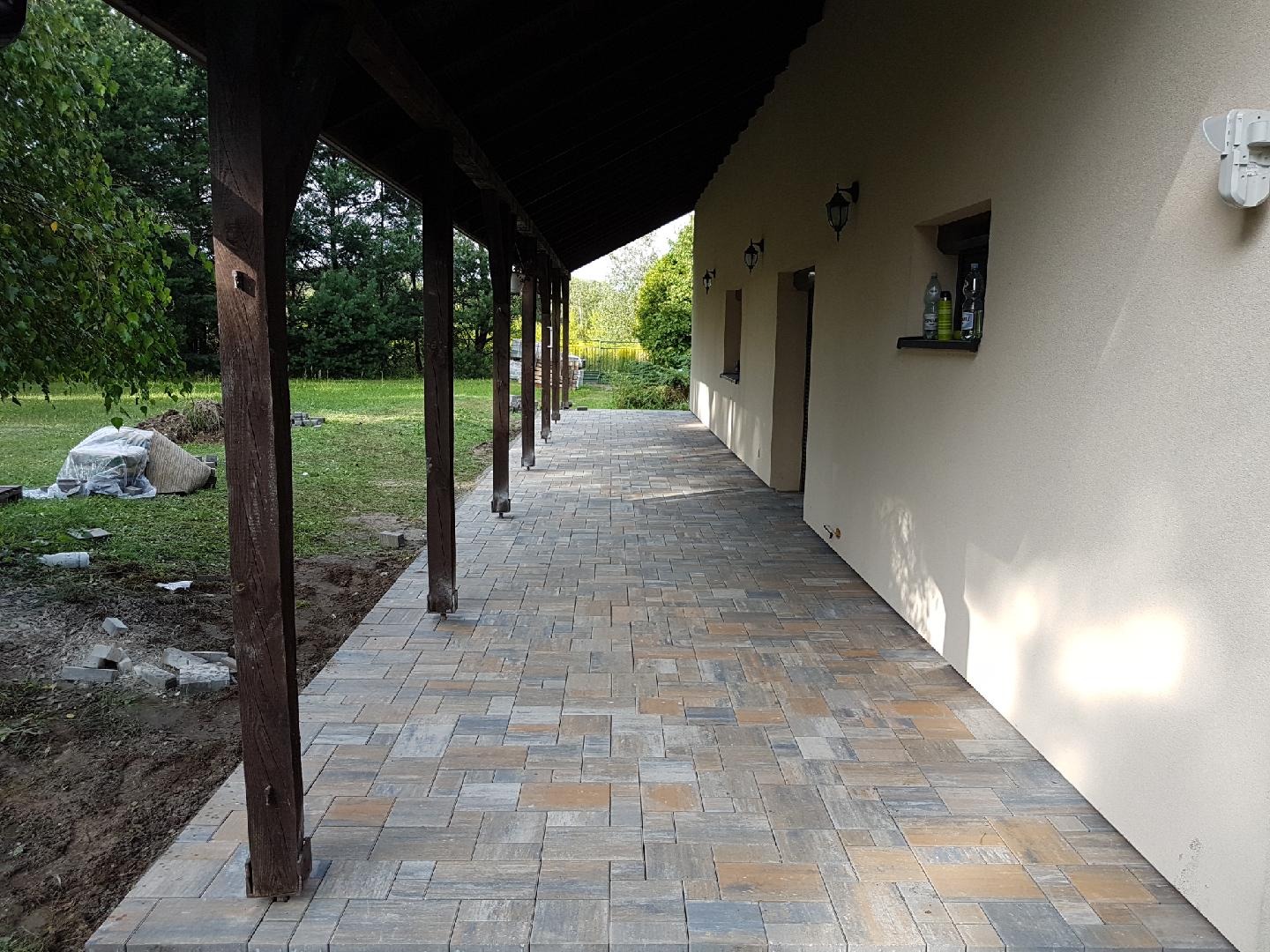 Wykończona ścieżka z kostki brukowej w odcieniach szarości i brązu, prowadząca wzdłuż ściany budynku z drewnianą kolumnadą, z widocznymi workami z materiałem budowlanym na trawniku.