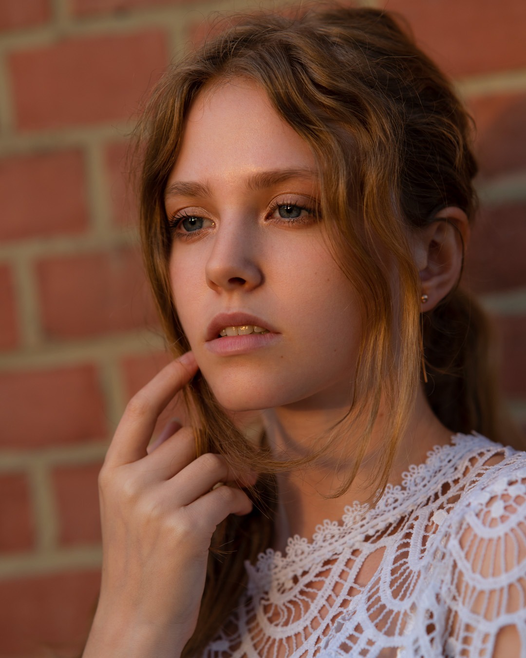 Portret młodej kobiety o jasnych włosach i niebieskich oczach, ubrana w białą, koronkową bluzkę, pozuje na tle ceglanej ściany w ciepłym, naturalnym świetle.