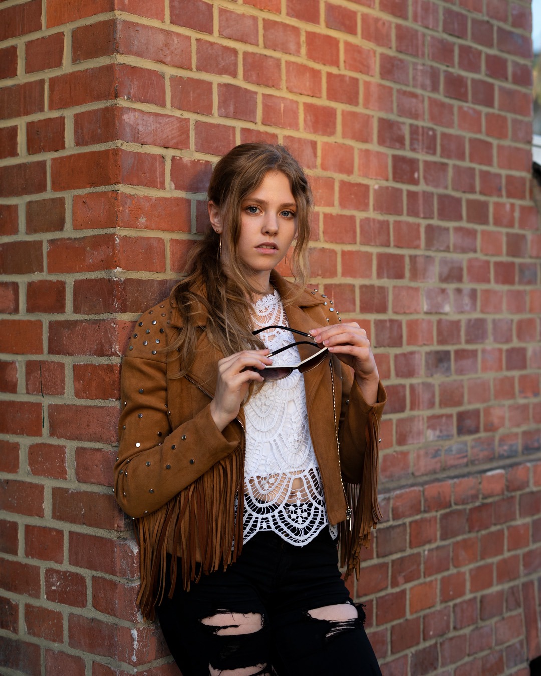 Młoda kobieta w kurtce z frędzlami i koronkowym topie pozuje oparta o ceglaną ścianę, trzymając okulary przeciwsłoneczne.
