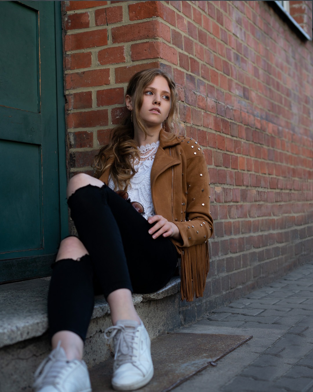 Młoda kobieta w brązowej kurtce z frędzlami i podartych dżinsach siedzi na schodach przy ceglanej ścianie, spoglądając w górę.
