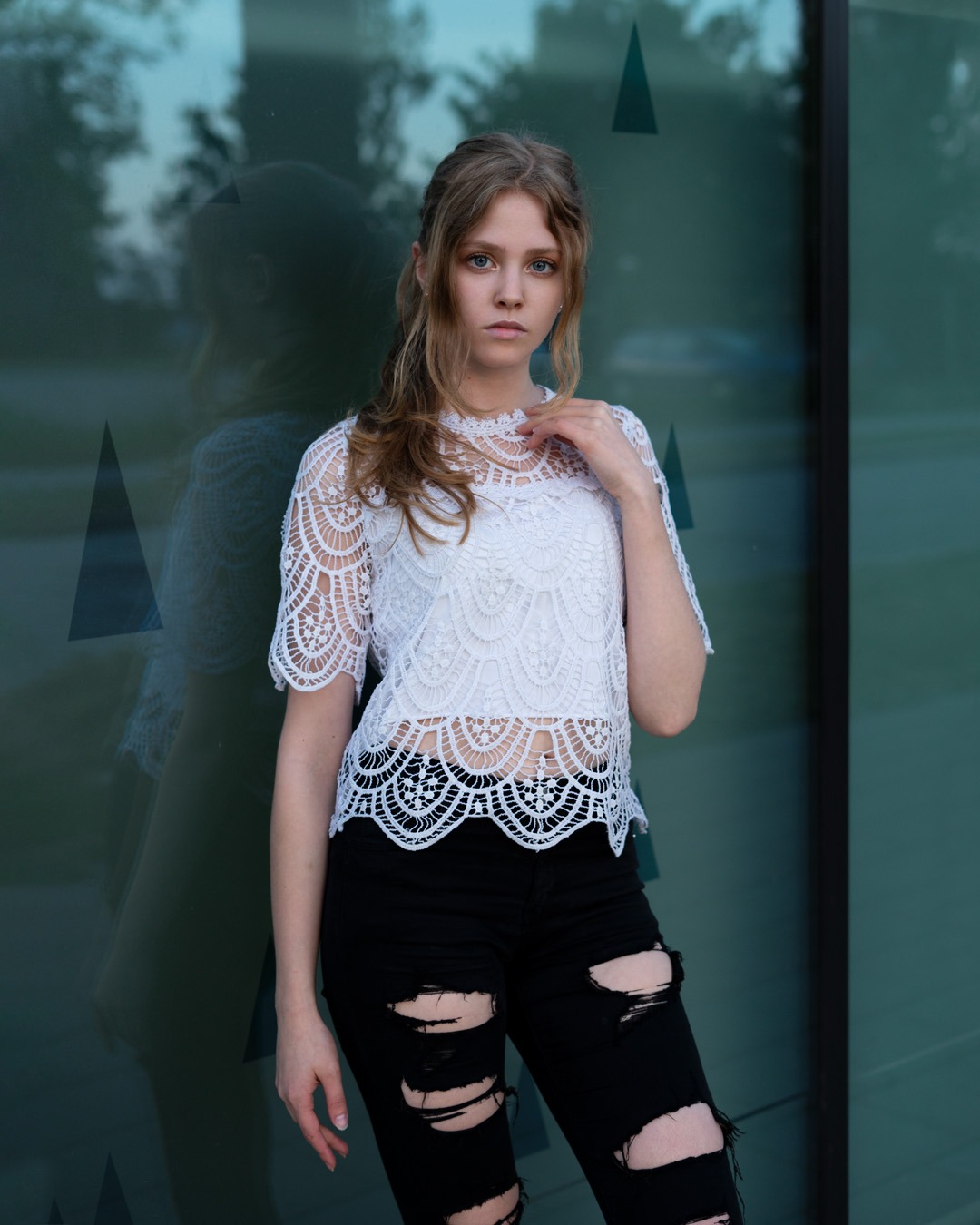 Młoda kobieta w koronkowej bluzce i podartych jeansach pozuje na tle szklanej ściany z widocznym odbiciem.