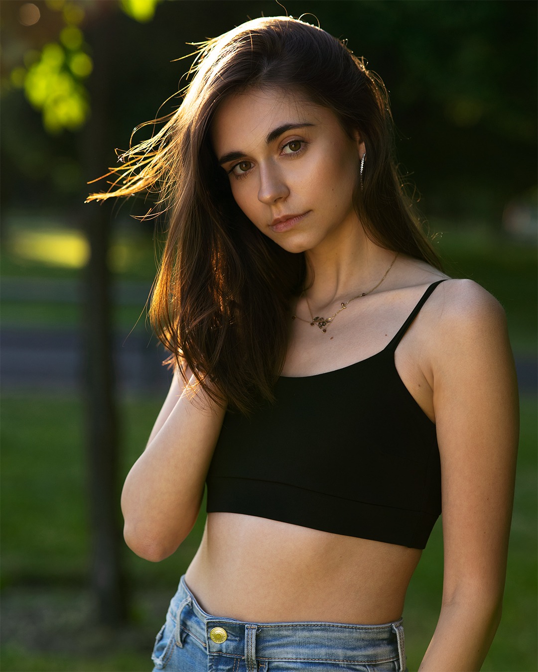 Portret młodej kobiety z długimi brązowymi włosami, w czarnym topie i jeansach, pozującej w plenerze z rozproszonym światłem słonecznym i zielonym tłem.
