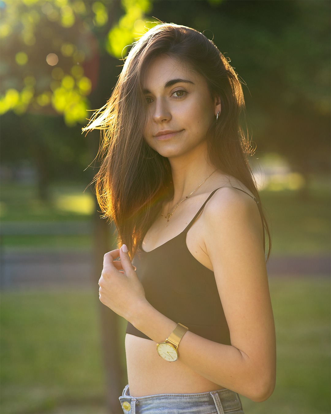 Portret młodej kobiety w letnim słońcu, z delikatnym uśmiechem, złotym zegarkiem i naszyjnikiem, na tle rozmytej zieleni.