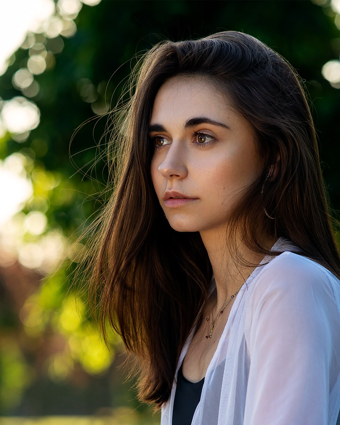 Portret młodej kobiety z długimi, brązowymi włosami, w zwiewnej, białej narzutce, spoglądającej w lewo na tle rozmytej zieleni i złotego światła.