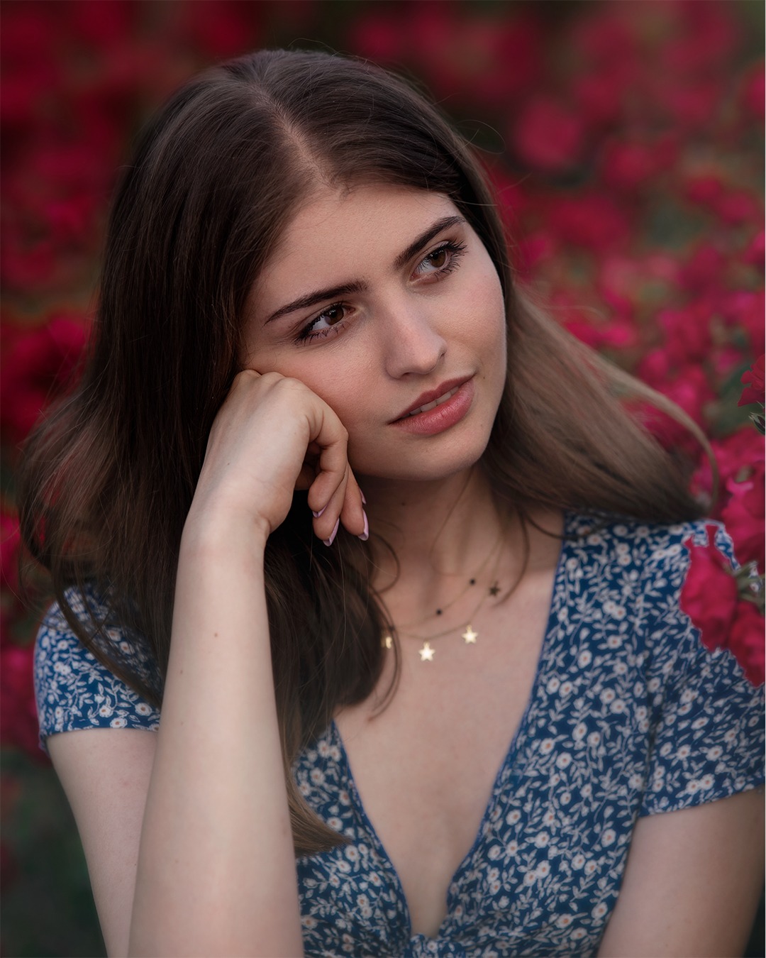 Portret młodej kobiety w kwiecistej sukience, opierającej policzek na dłoni, z delikatnym naszyjnikiem w kształcie gwiazdek na tle intensywnie czerwonych kwiatów.