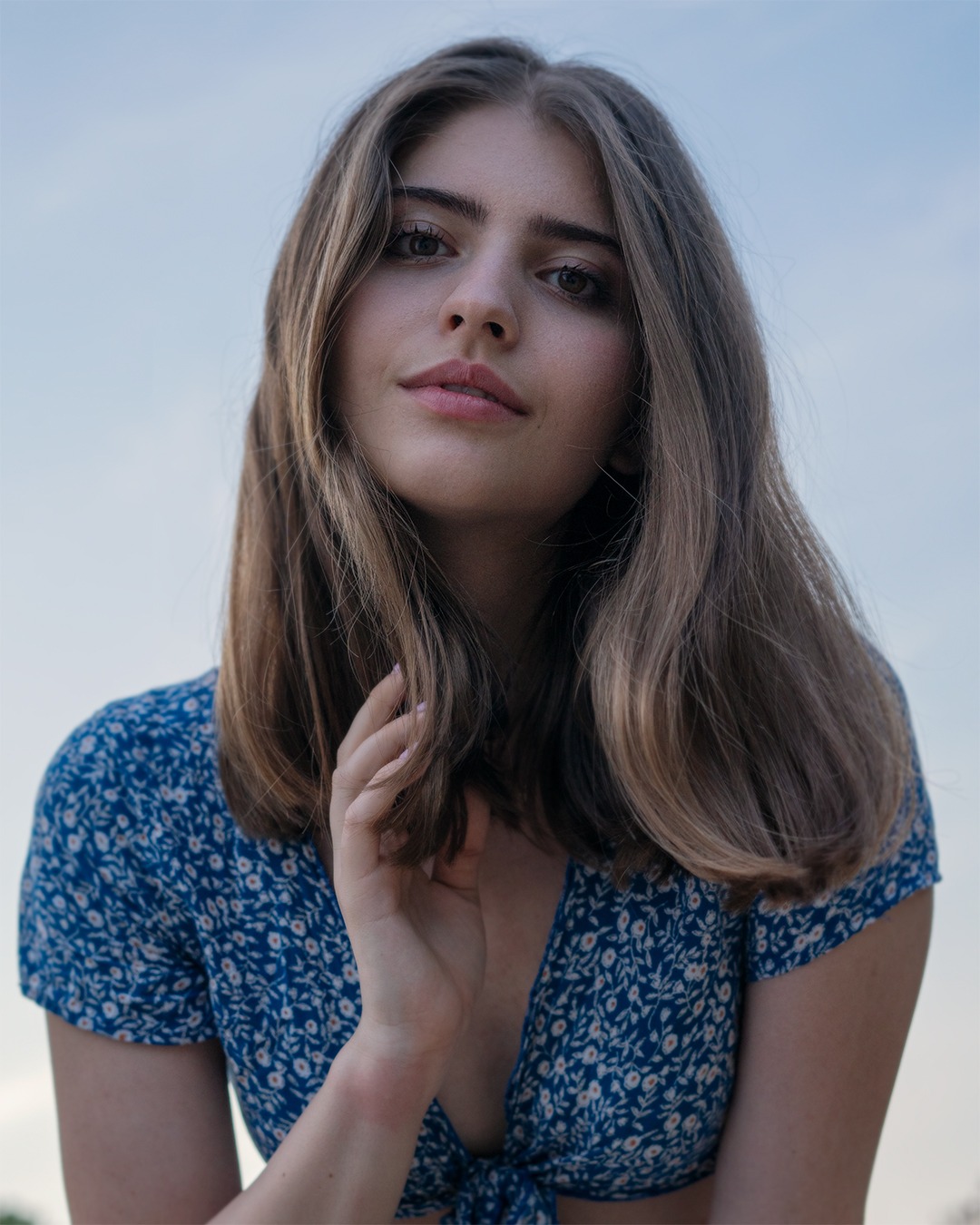 Portret młodej kobiety w niebieskiej sukience w kwiaty, z ręką delikatnie dotykającą szyi, patrzącej w górę na fotografa na tle jasnego nieba.