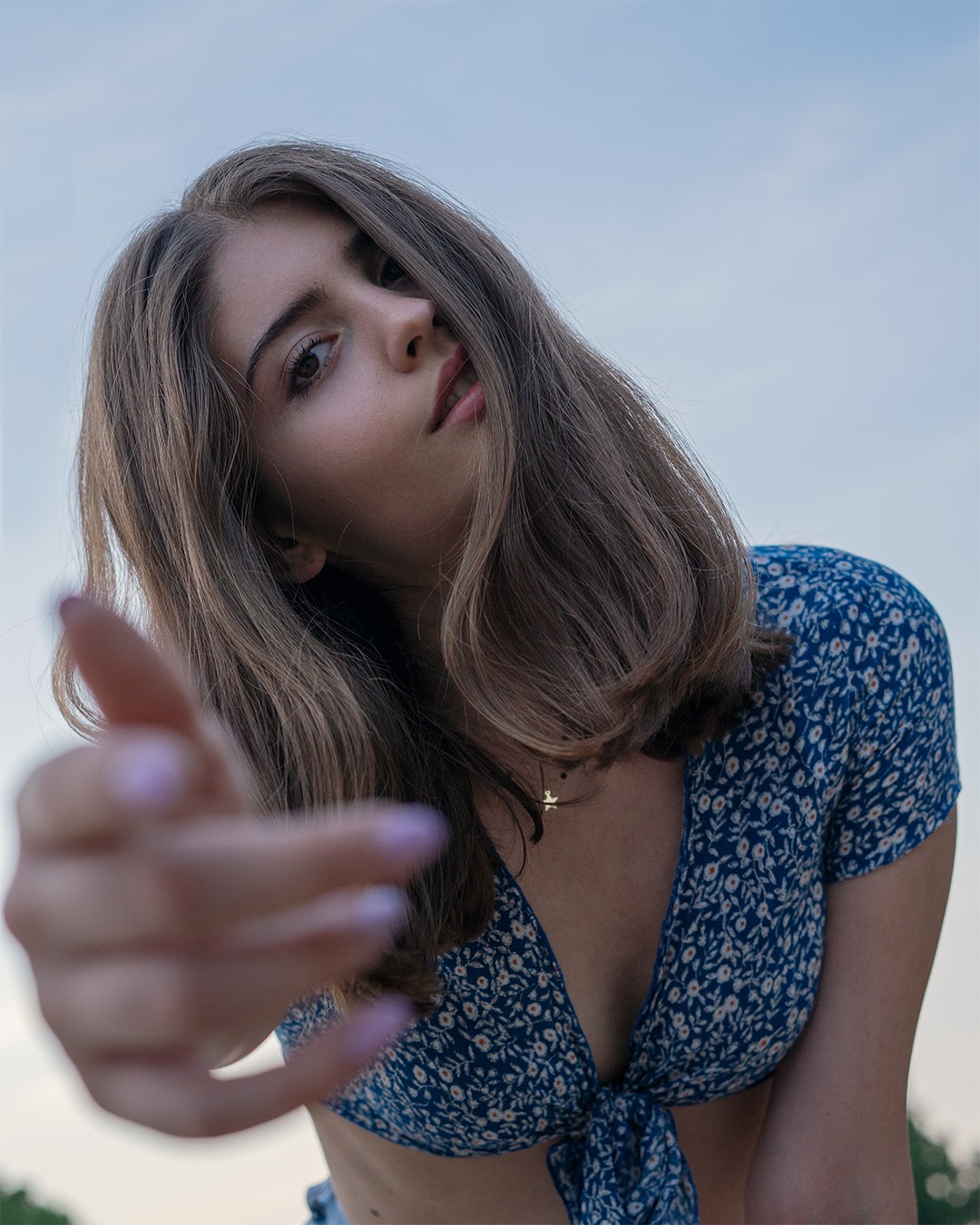 Portret młodej kobiety w kwiecistej bluzce, z wyciągniętą ręką w kierunku obiektywu, sfotografowana z dołu na tle jasnego nieba.