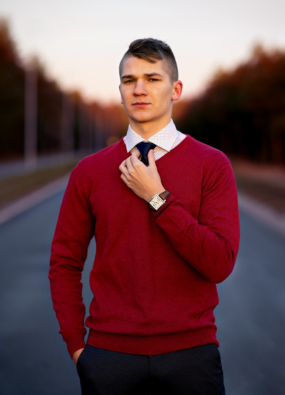 Młody mężczyzna w bordowym swetrze i koszuli w kropki, poprawiający krawat na tle rozmytej drogi i drzew w jesiennych kolorach.