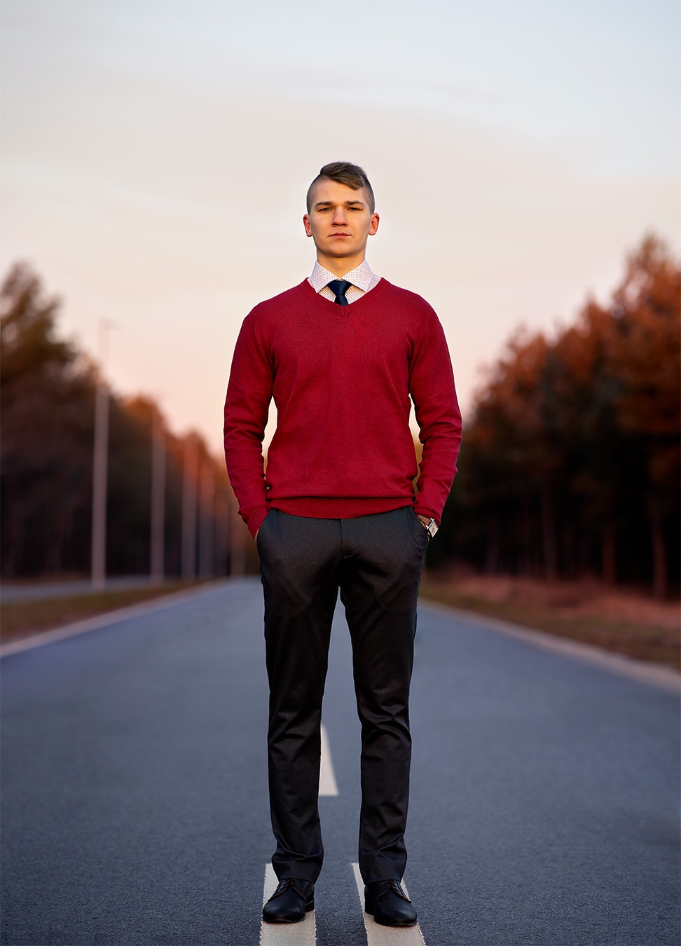 Młody mężczyzna w bordowym swetrze, białej koszuli i krawacie, stojący na środku asfaltowej drogi z rękami w kieszeniach, w tle drzewa i zachodzące słońce.