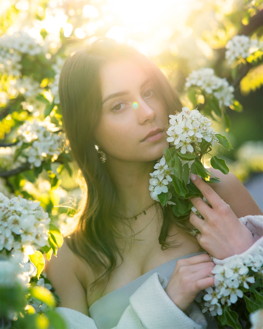 Portret młodej kobiety w kwitnącym sadzie, z gałązką białych kwiatów w dłoni, delikatne światło słoneczne, zwiewna sukienka.