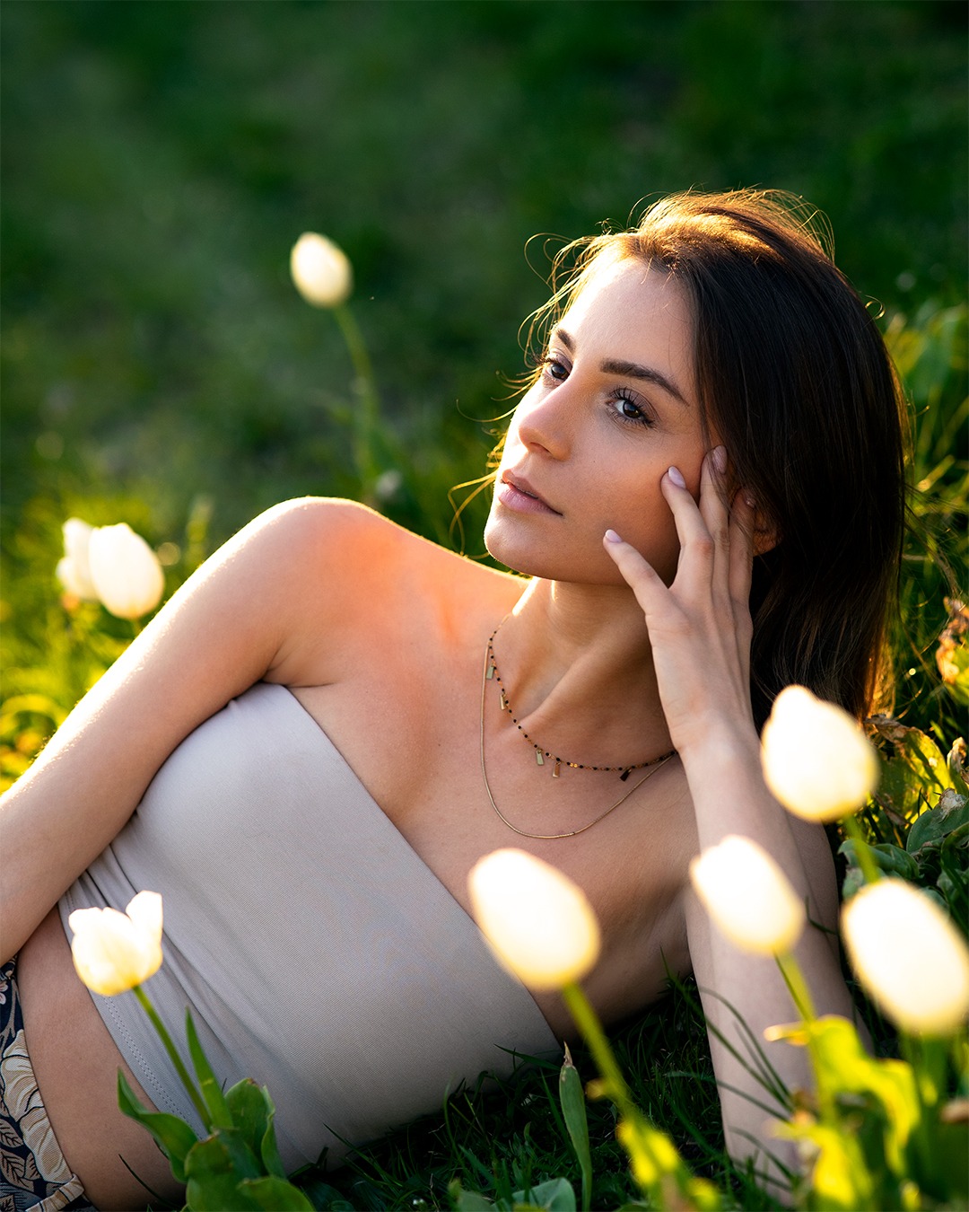 Kobieta leży na trawie wśród białych tulipanów, z delikatnym naszyjnikiem i ręką opartą o policzek, spoglądając w górę w miękkim świetle.