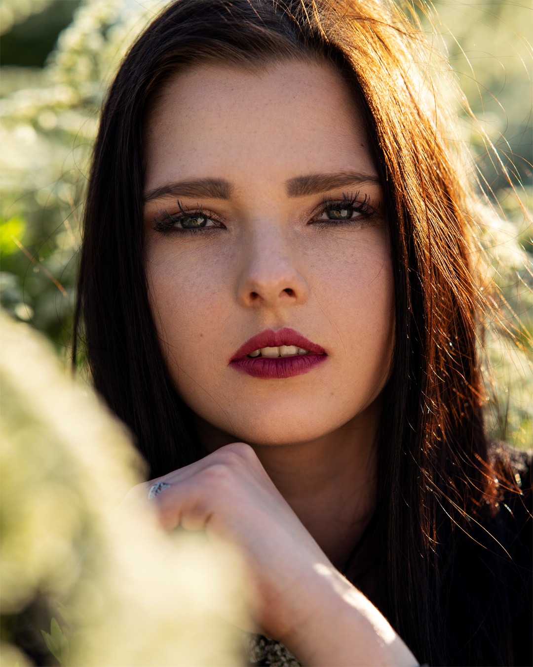 Portret kobiety z ciemnymi włosami i bordowymi ustami, opierającej podbródek na dłoni z pierścionkiem, na tle rozmytych białych kwiatów i promieni słońca.