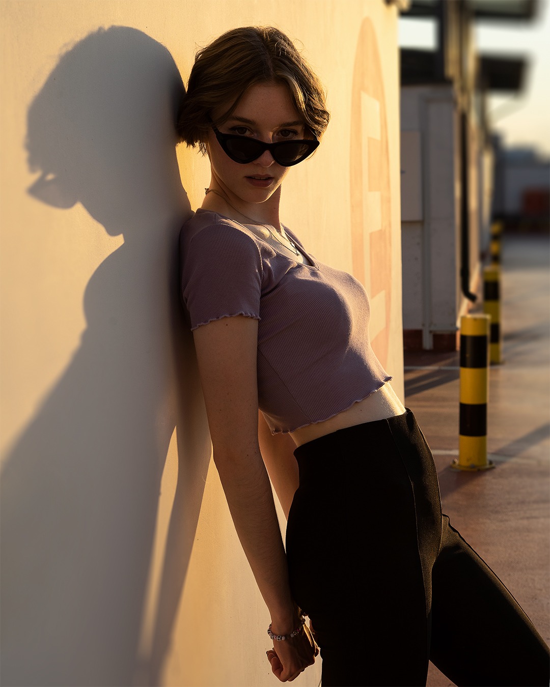 Młoda kobieta w okularach przeciwsłonecznych, oparta o ścianę w ciepłym, popołudniowym świetle, z wyraźnym cieniem na ścianie i widocznym fragmentem parkingu w tle.