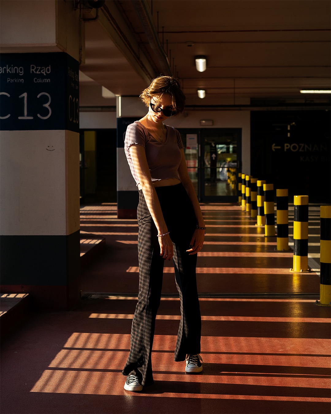 Kobieta w okularach przeciwsłonecznych pozuje w garażu, oświetlona promieniami słońca, w spodniach w kratę i krótkiej bluzce.