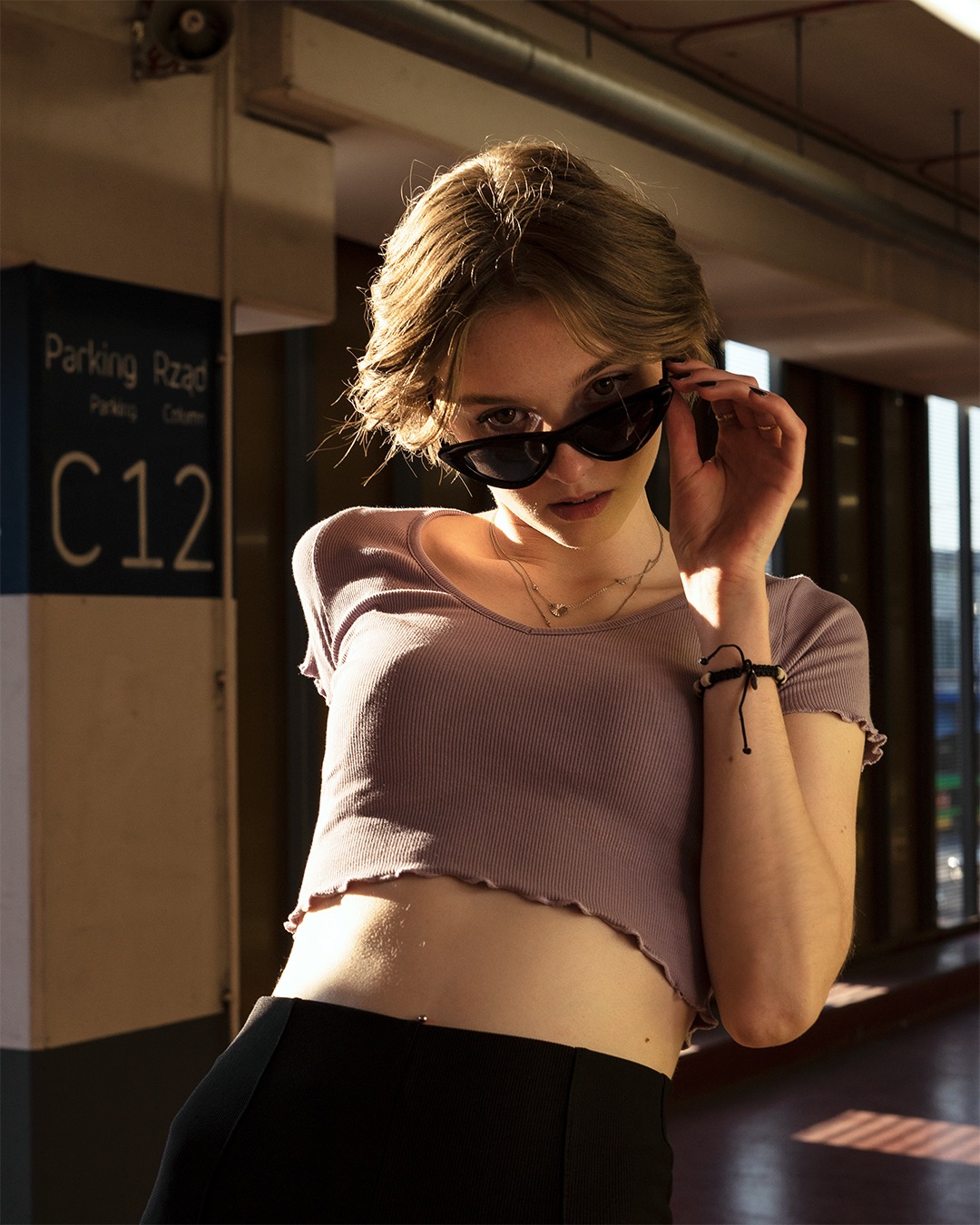 Portret młodej kobiety w okularach przeciwsłonecznych, pozującej w garażu podziemnym, z widocznym znakiem 'Parking Rząd C12'.