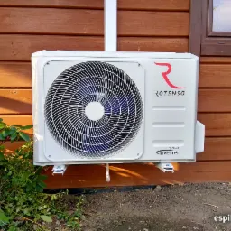Biała jednostka zewnętrzna klimatyzacji Rotenso Inverter zamontowana na drewnianej elewacji domu, widoczna z boku krzewu i glinianego dzbanka.