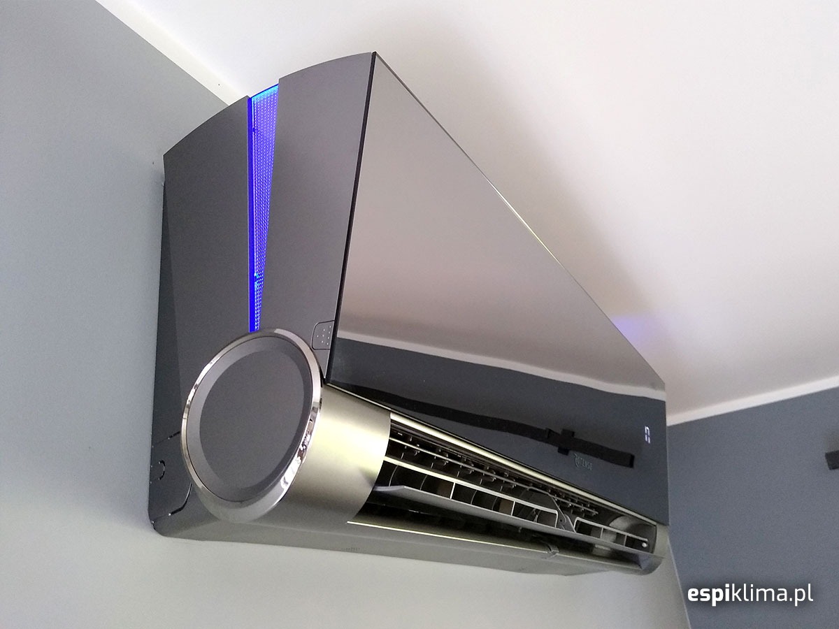 Nowoczesny, szary klimatyzator ścienny z chromowanymi detalami i niebieskim podświetleniem LED, zamontowany w rogu pomieszczenia z szarą ścianą.