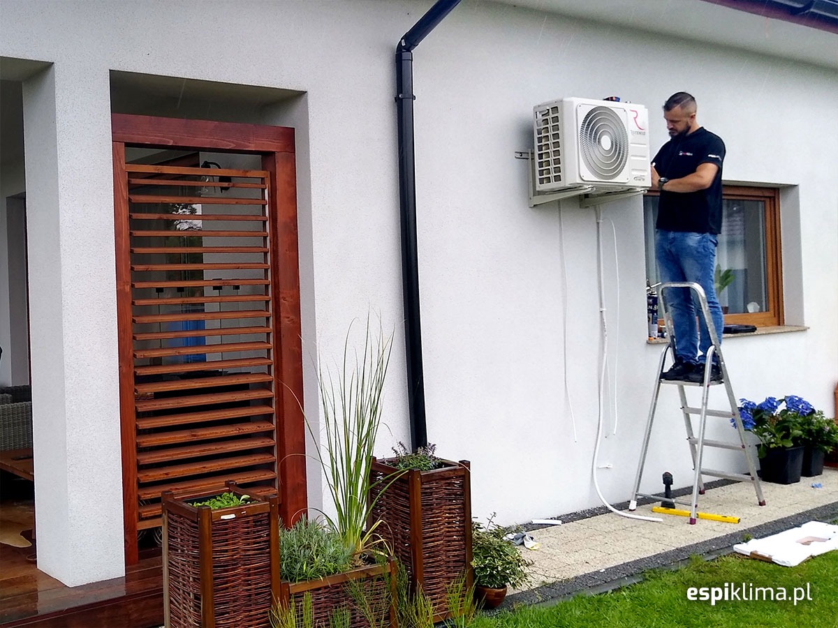 Mężczyzna na drabinie instaluje klimatyzator na ścianie domu, widoczne przewody i elementy montażowe, obok okno i dekoracyjne donice z roślinami.