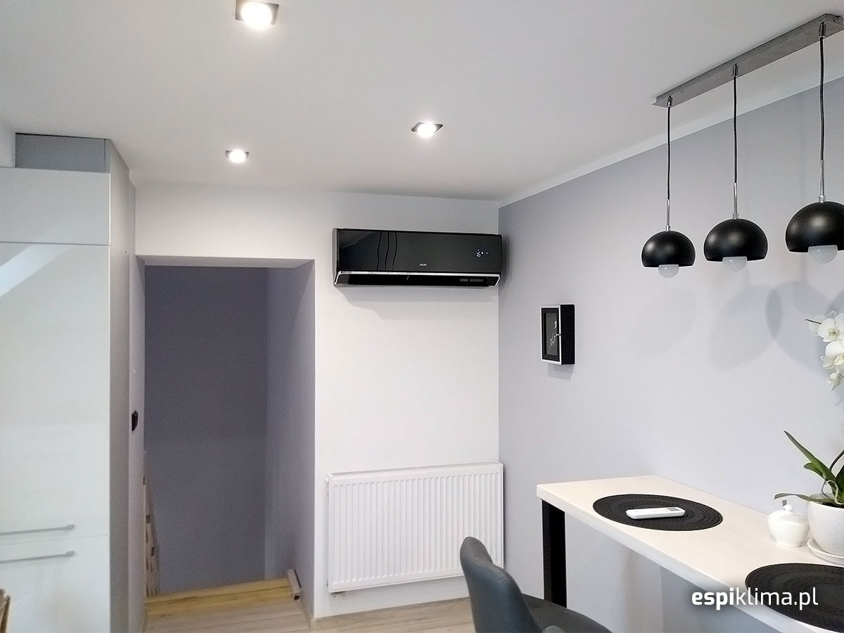 Czarny klimatyzator ścienny w nowoczesnym wnętrzu z białymi ścianami, grzejnikiem i minimalistycznym oświetleniem.