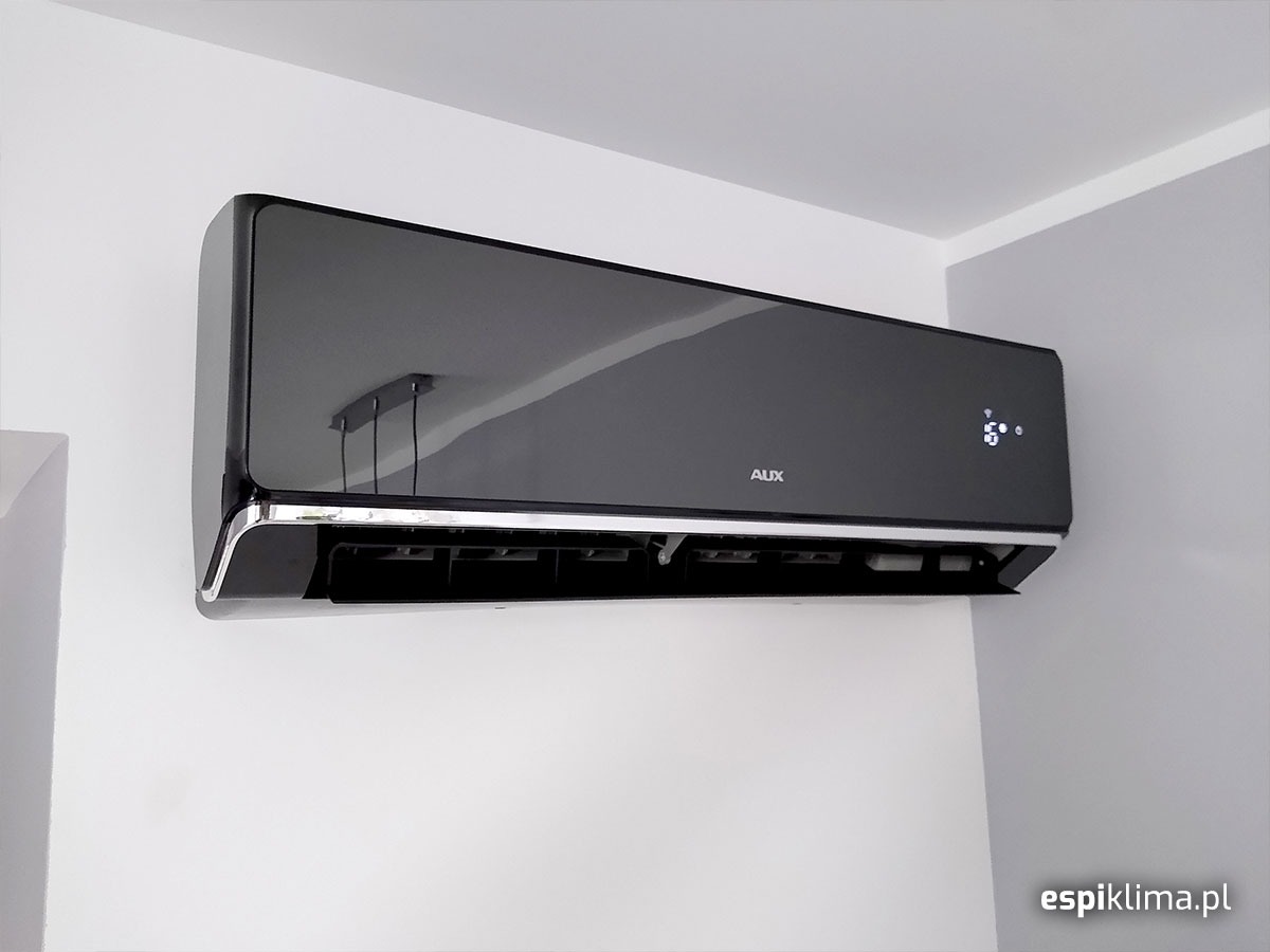 Czarny, błyszczący klimatyzator marki AUX zamontowany na szarej ścianie w rogu pokoju, z widocznym odbiciem lampy na obudowie.