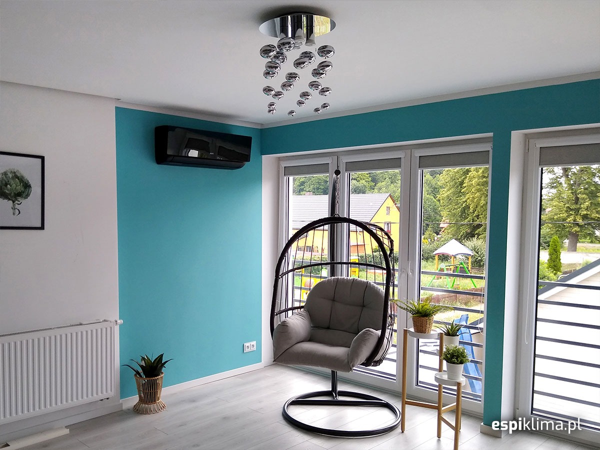 Nowoczesny salon z czarnym klimatyzatorem ściennym, wiszącym fotelem typu kokon i widokiem na plac zabaw przez okno.
