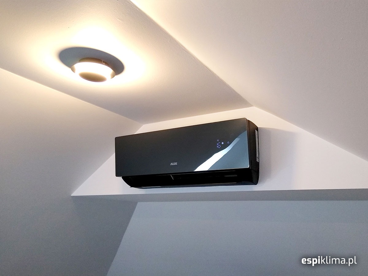 Czarny klimatyzator AUX zamontowany w rogu pomieszczenia pod skosem sufitu, widoczny wyświetlacz z temperaturą 16 stopni.