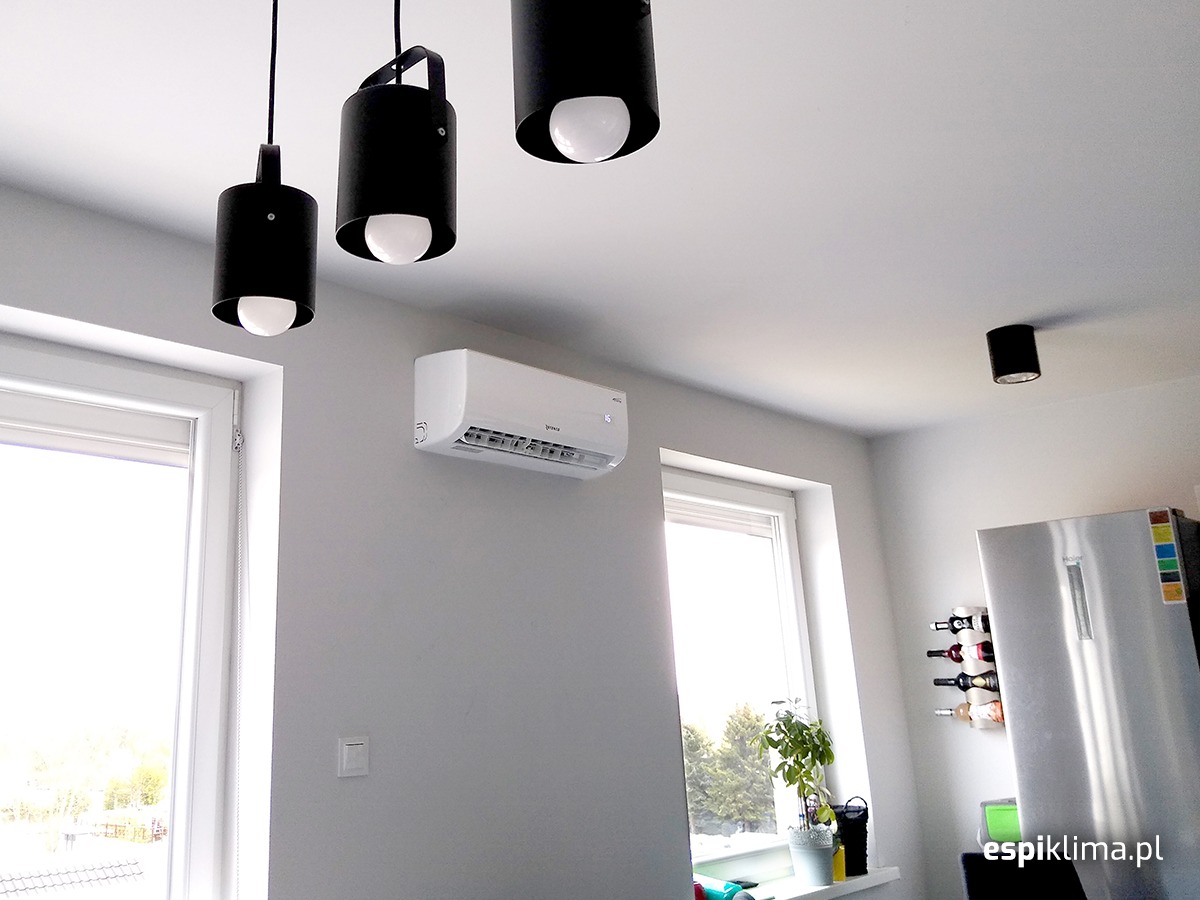 Biały klimatyzator zamontowany na szarej ścianie w jasnym, nowoczesnym wnętrzu mieszkania, widoczne trzy czarne lampy wiszące i okno.