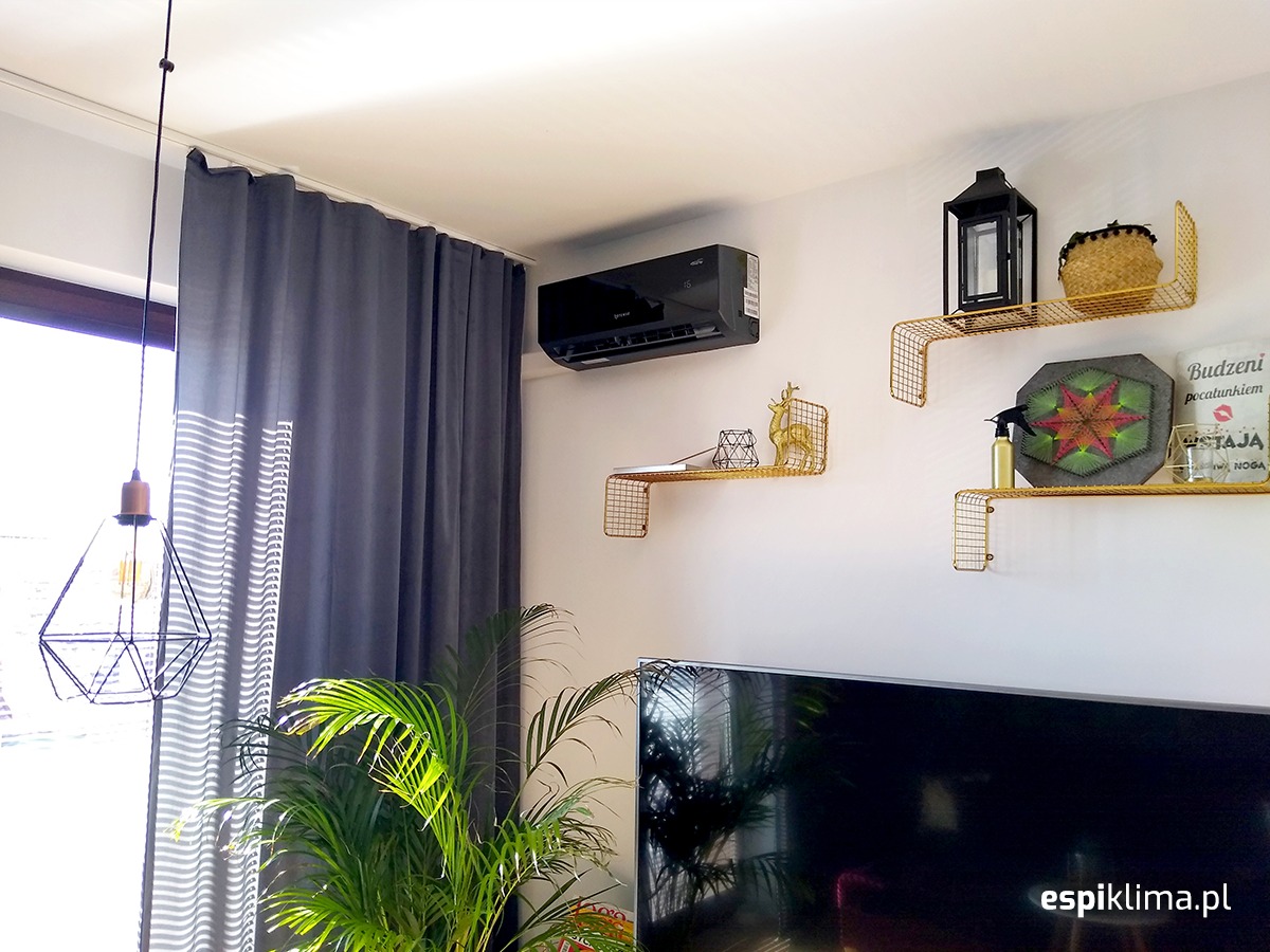 Czarny klimatyzator ścienny w nowoczesnym salonie z dekoracyjnymi złotymi półkami, zasłonami i rośliną doniczkową.