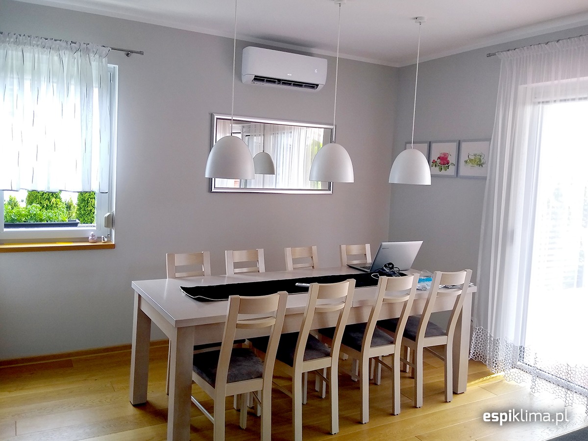 Biały klimatyzator ścienny zamontowany nad lustrem w minimalistycznej jadalni z długim stołem, laptopem i trzema lampami wiszącymi.