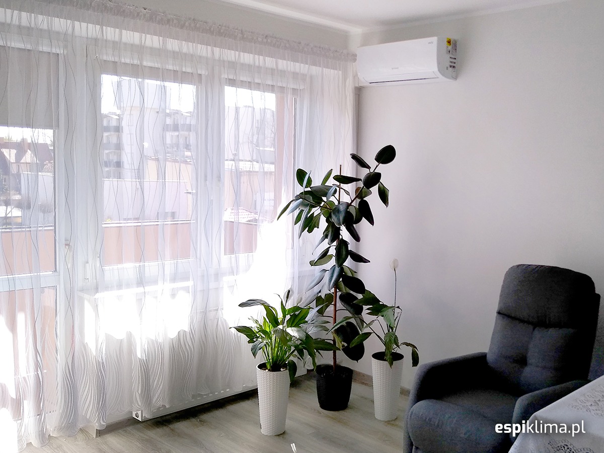 Biały klimatyzator na ścianie w jasnym pokoju z dużym oknem zasłoniętym firankami, obok rośliny doniczkowe i szary fotel.