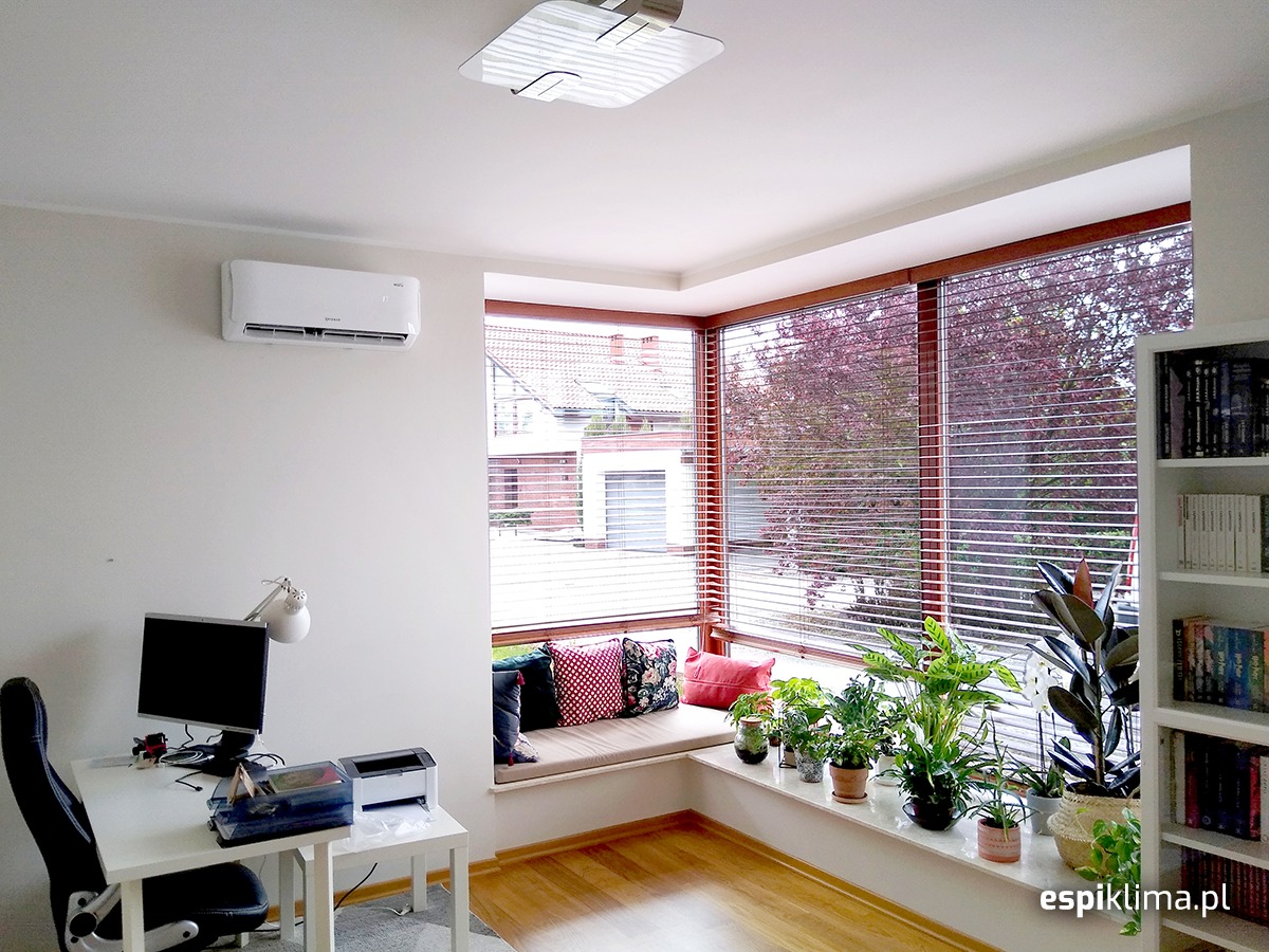 Biały klimatyzator ścienny zamontowany w jasnym pokoju z dużym oknem, parapetem pełnym roślin doniczkowych i biurkiem z komputerem.