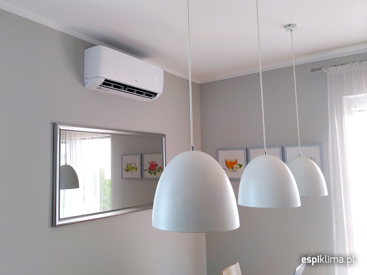 Biały klimatyzator ścienny zamontowany w pokoju z szarymi ścianami, pod nim lustro w srebrnej ramie, w tle trzy białe lampy wiszące i okno z białymi zasłonami.