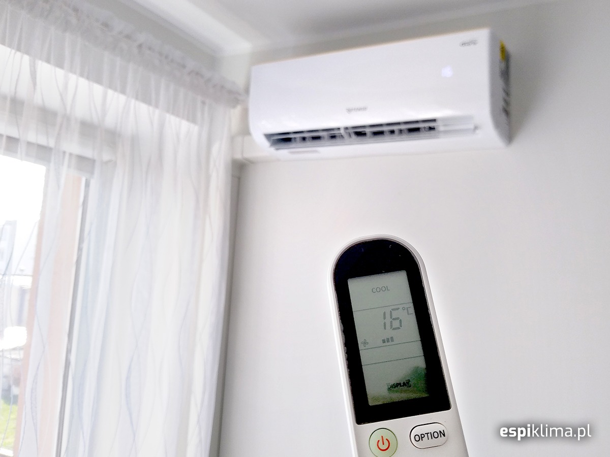 Biały klimatyzator ścienny z pilotem ustawionym na 16 stopni Celsjusza w jasnym pomieszczeniu w Lubinie, widoczne zasłony w oknie.