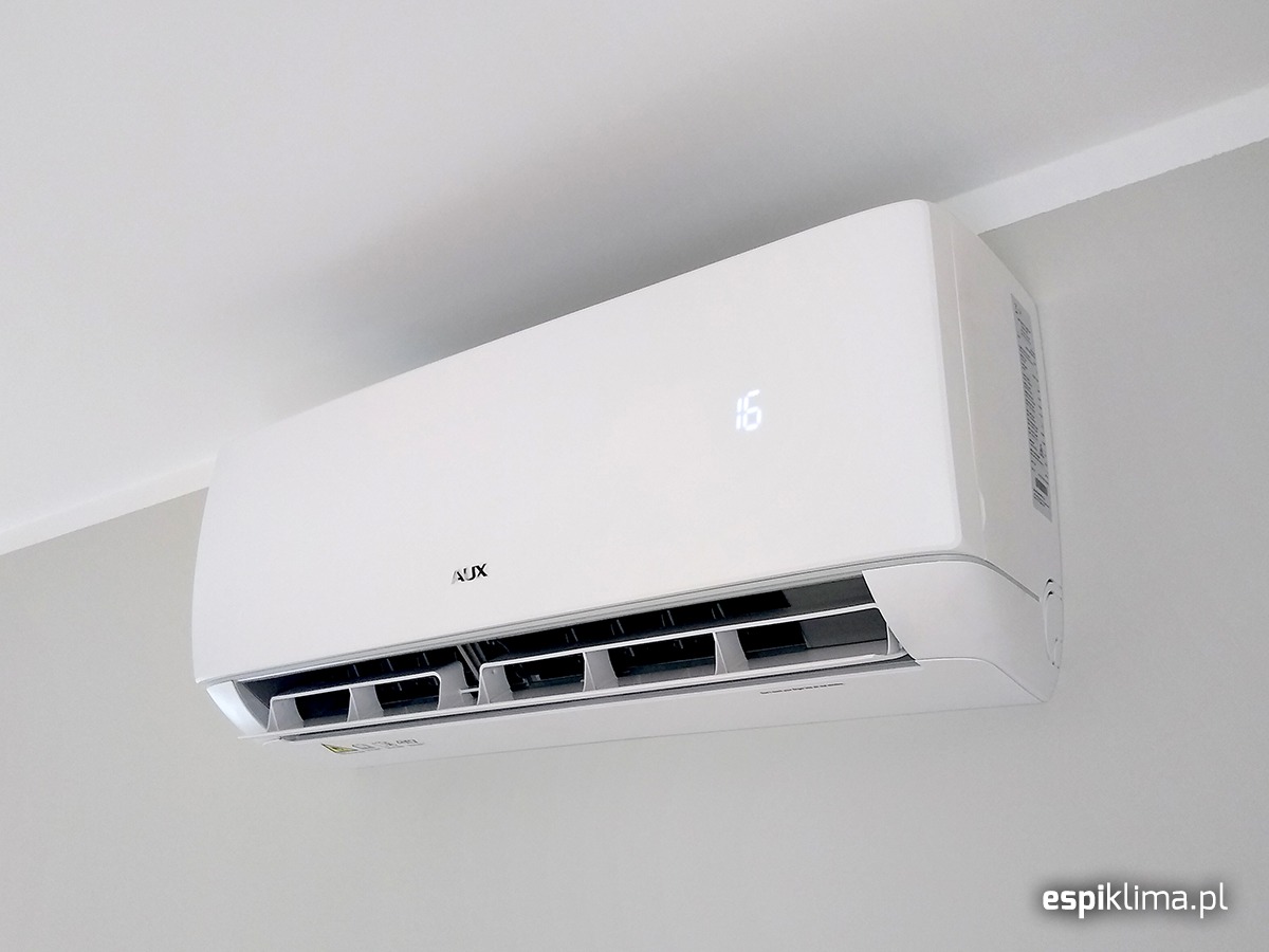 Biały klimatyzator AUX zamontowany na ścianie, wyświetlacz pokazuje temperaturę 16 stopni Celsjusza.