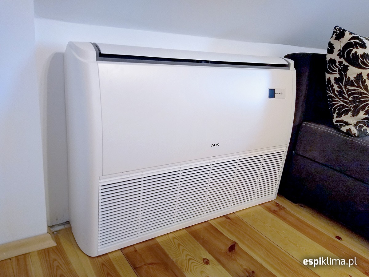Biały klimatyzator podłogowy marki AUX ustawiony przy ścianie w pomieszczeniu z drewnianą podłogą, obok szarej sofy z ozdobną poduszką.