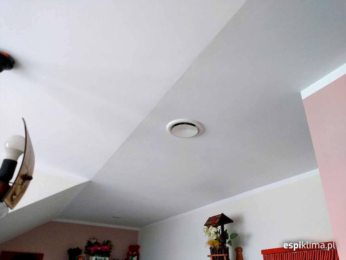 Okrągły nawiewnik sufitowy klimatyzacji w pomieszczeniu mieszkalnym, widoczny pod kątem od dołu, z elementami dekoracyjnymi w tle i lampą po lewej stronie.
