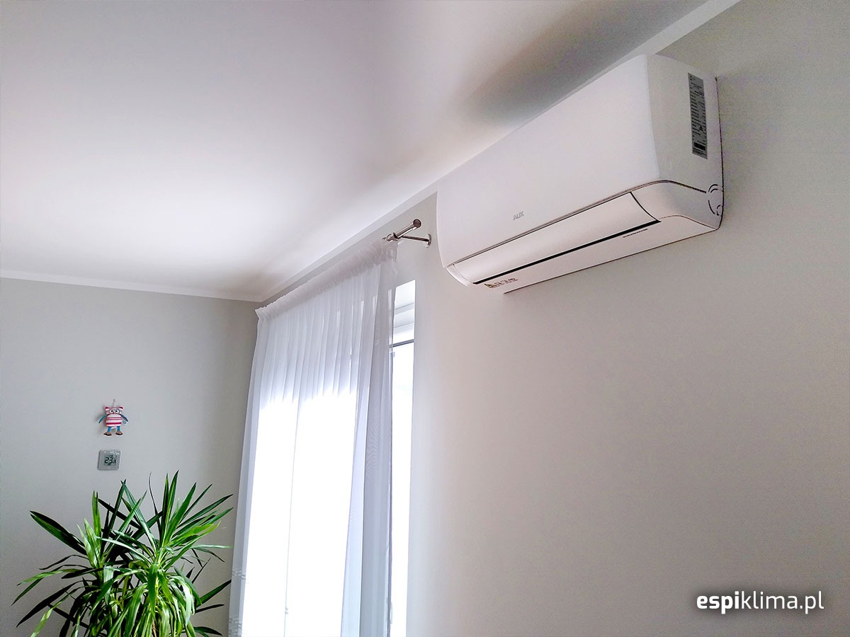 Biały klimatyzator AUX zamontowany na jasnoszarej ścianie w pokoju, widok z dołu, obok okna z białymi zasłonami i zielonej rośliny doniczkowej.