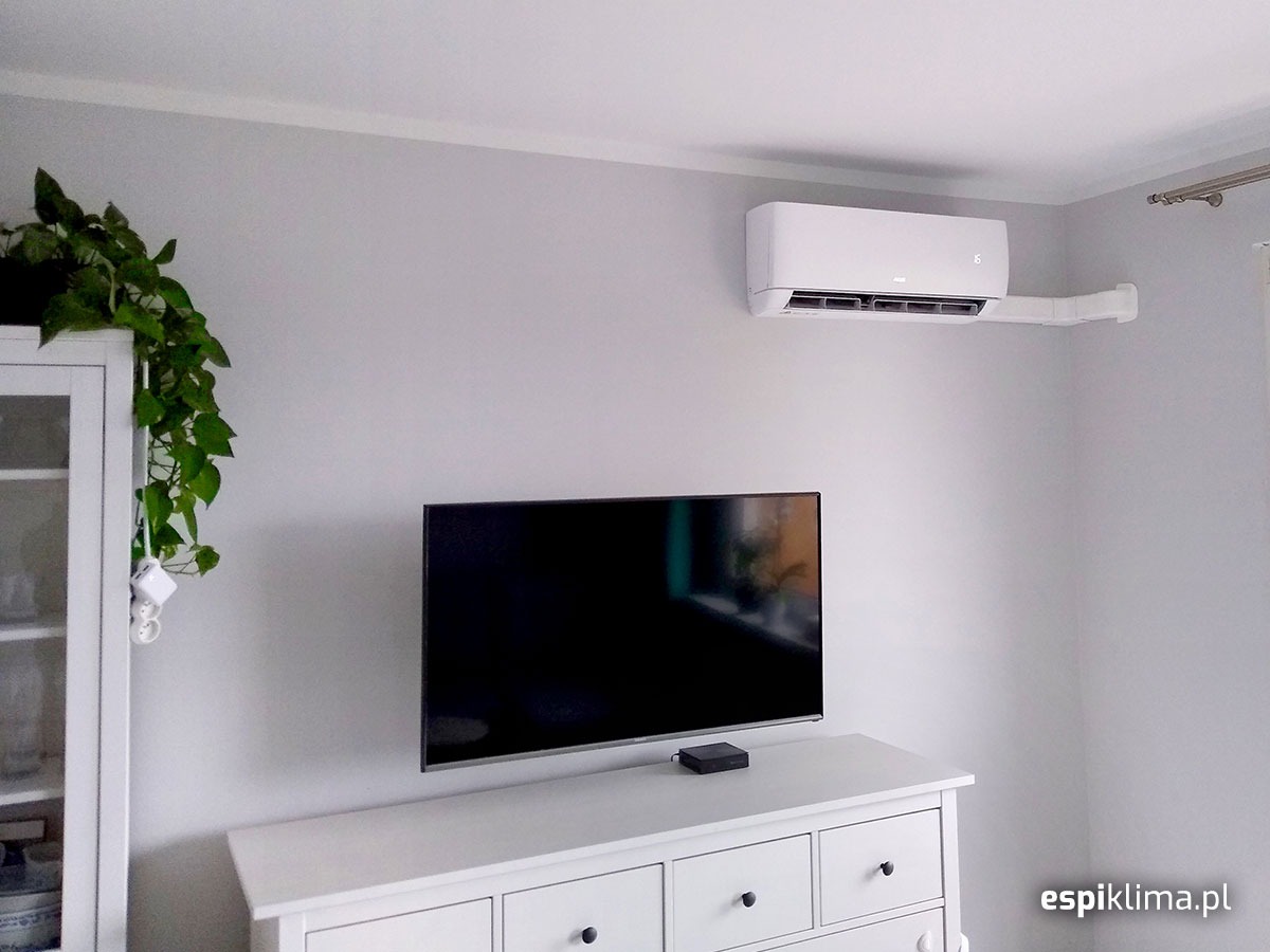 Biały klimatyzator ścienny zamontowany w pokoju nad telewizorem, z widoczną białą listwą przypodłogową i szafką z rośliną doniczkową w tle.