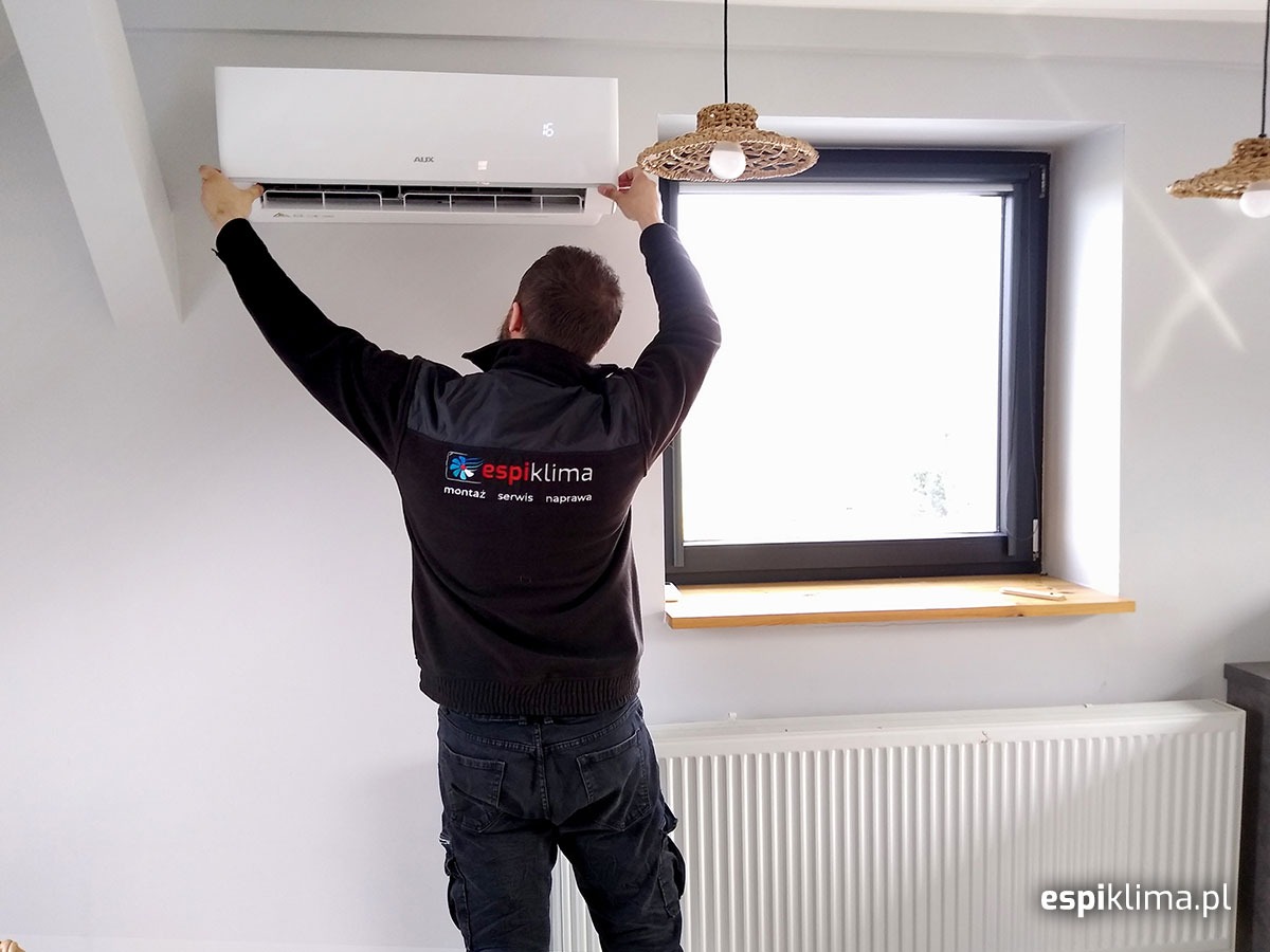 Mężczyzna w firmowej bluzie montuje klimatyzator AUX na białej ścianie w pomieszczeniu z grzejnikiem i oknem, widoczne lampy z abażurami z rafii.