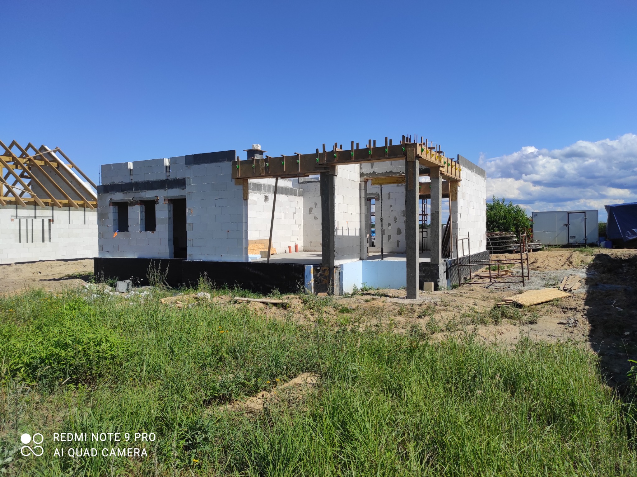 Wznoszenie ścian domu z białych bloczków, widoczne szalunki stropowe, w tle konstrukcja dachu drugiego budynku i kontener budowlany.