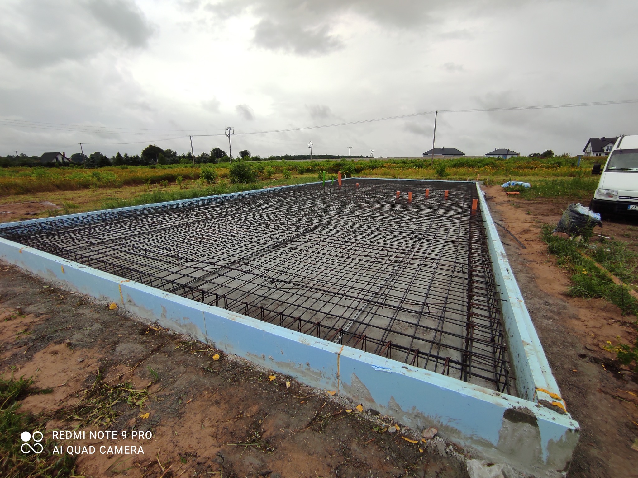Zbrojenie fundamentu domu jednorodzinnego z użyciem prętów stalowych i szalunków z izolacją termiczną, widoczne wystające rury instalacyjne, tło stanowią pola i zabudowa jednorodzinna...
