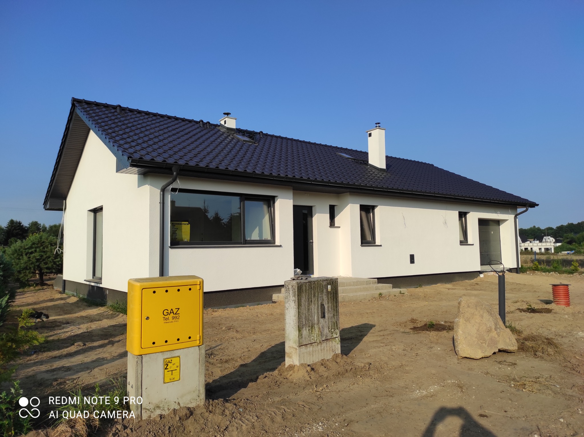 Nowoczesny dom jednorodzinny z białymi ścianami i ciemną dachówką w Koszalinie, widok z zewnątrz na tle błękitnego nieba, z żółtą skrzynką gazową na pierwszym planie.