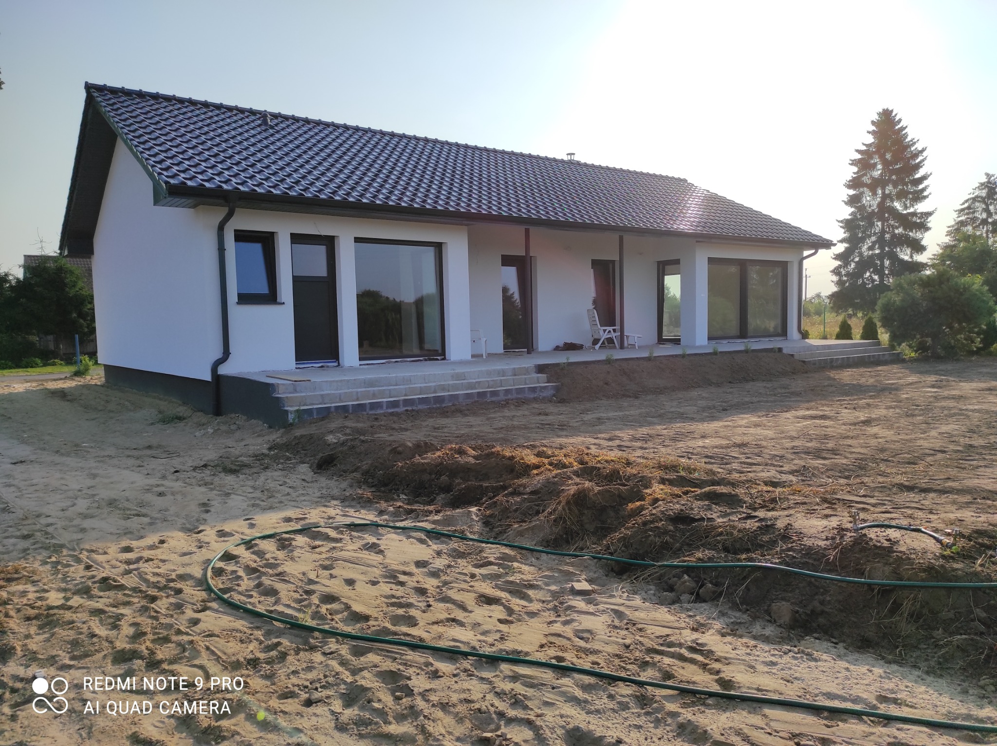 Nowoczesny, parterowy dom z białymi ścianami i ciemną dachówką, widoczny z zewnątrz na działce w trakcie zagospodarowywania, z widocznymi schodami wejściowymi i zielonym wężem ogrodowym...